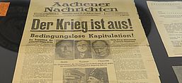 Titelseite der Zeitung Aachener Nachrichten vom 8. Mai 1945. Die Hauptüberschrift in großen, fetten Lettern lautet: „Der Krieg ist aus!“. Darunter steht: „Bedingungslose Kapitulation!“ Die Zeitung berichtet vom Ende des Zweiten Weltkriegs in Europa. In der Mitte sind Porträts von Stalin, Truman, Churchill, Hitler und Mussolini abgedruckt. Weitere Artikel thematisieren unter anderem die Kapitulation der deutschen Wehrmacht und den Beginn des Friedens in Europa. Die Zeitung liegt in einer Ausstellungsvitrine, daneben sind weitere historische Dokumente zu sehen.