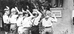 Das schwarz-weiße Foto zeigt eine Gruppe von Jungen in kurzen Hosen, Hemden und Halstüchern, die vor einer Statue mit der Aufschrift „Der kleine Trompeter“ salutieren. Die Statue stellt eine männliche Figur in Uniform dar, die ein Musikinstrument in der Hand hält. Sie steht auf einem steinernen Sockel. Die Szenerie befindet sich in einer bewaldeten, felsigen Umgebung mit mehreren Erwachsenen im Hintergrund, die das Geschehen beobachten. Junge Pioniere aus der DDR-Zeit nehmen an einer Gedenkveranstaltung teil.