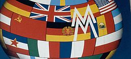 Ausschnitt aus dem Plakat der Leipziger Messe (1989): Eine kugelförmige Oberfläche aus aneinandergesetzten Nationalflaggen füllt das Bild. Über die Flaggen legt sich ein weißes, stilisiertes „M“ als Messezeichen; der Hintergrund ist blau.