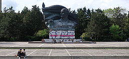 Das Bild zeigt das Ernst-Thälmann-Denkmal in Berlin, eine monumentale Bronzeskulptur, die Ernst Thälmann, den Vorsitzenden der Kommunistischen Partei Deutschlands (KPD) in den 1920er und 1930er Jahren, darstellt. Die Skulptur zeigt Thälmann mit erhobener Faust, ein Symbol für den kommunistischen Kampf. Im unteren Bereich des Denkmals ist der Name "Ernst Thälmann" in großen Buchstaben zu sehen, allerdings ist die Basis des Denkmals mit Graffiti und Farbmarkierungen übersät. Vor dem Denkmal sitzen zwei Personen auf den Stufen, während sich Fahrräder und andere Menschen im Hintergrund befinden. Im Hintergrund ist eine dichte Baumreihe zu sehen.