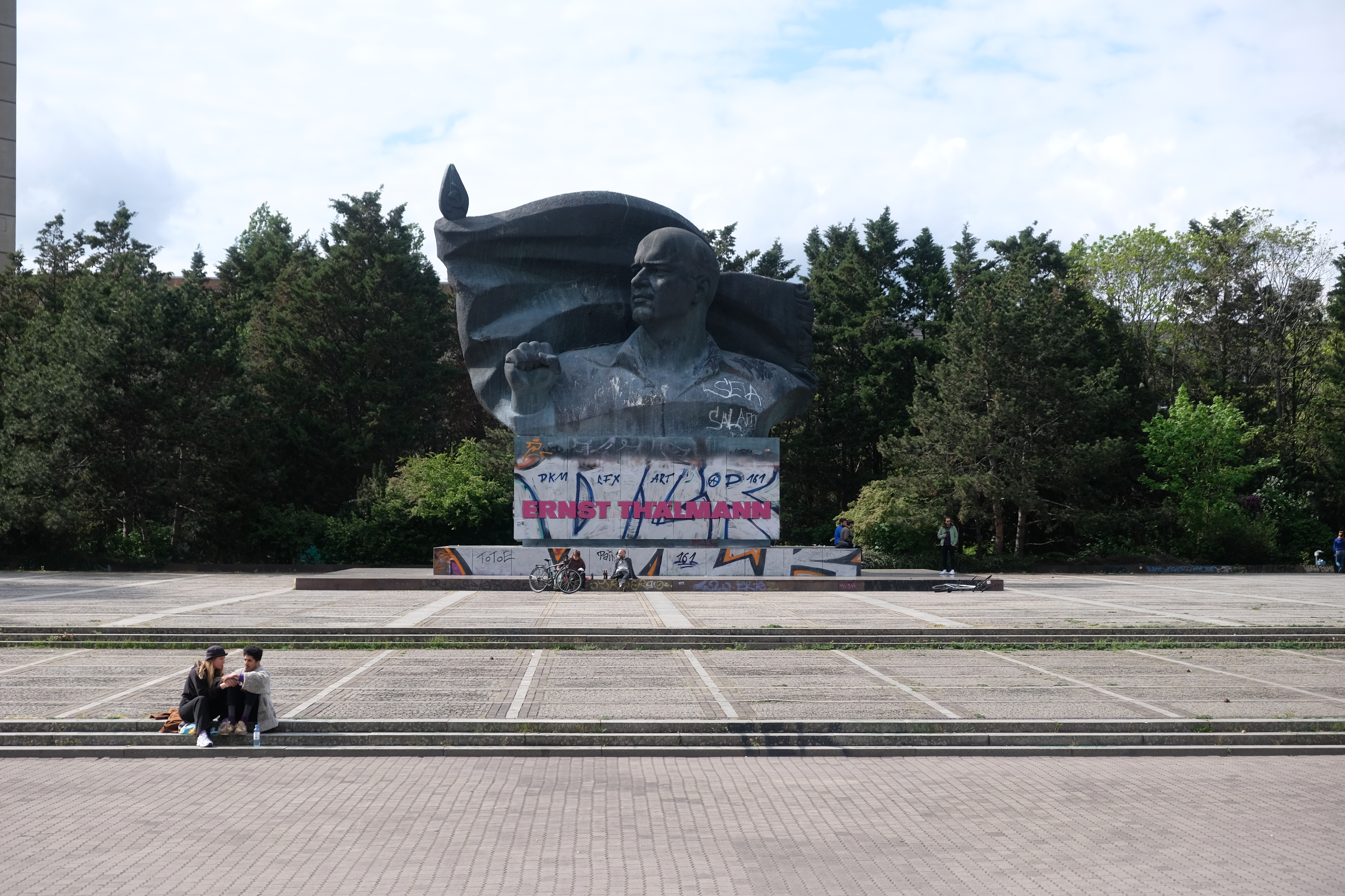 Das Bild zeigt das Ernst-Thälmann-Denkmal in Berlin, eine monumentale Bronzeskulptur, die Ernst Thälmann, den Vorsitzenden der Kommunistischen Partei Deutschlands (KPD) in den 1920er und 1930er Jahren, darstellt. Die Skulptur zeigt Thälmann mit erhobener Faust, ein Symbol für den kommunistischen Kampf. Im unteren Bereich des Denkmals ist der Name "Ernst Thälmann" in großen Buchstaben zu sehen, allerdings ist die Basis des Denkmals mit Graffiti und Farbmarkierungen übersät. Vor dem Denkmal sitzen zwei Personen auf den Stufen, während sich Fahrräder und andere Menschen im Hintergrund befinden. Im Hintergrund ist eine dichte Baumreihe zu sehen.