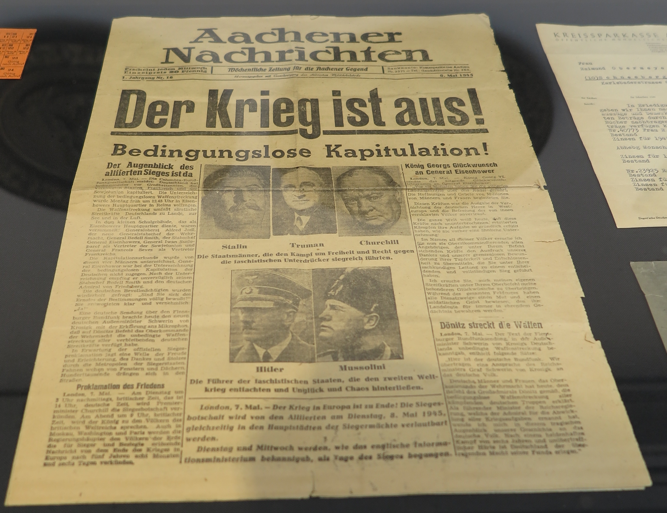 Titelseite der Zeitung Aachener Nachrichten vom 8. Mai 1945. Die Hauptüberschrift in großen, fetten Lettern lautet: „Der Krieg ist aus!“. Darunter steht: „Bedingungslose Kapitulation!“ Die Zeitung berichtet vom Ende des Zweiten Weltkriegs in Europa. In der Mitte sind Porträts von Stalin, Truman, Churchill, Hitler und Mussolini abgedruckt. Weitere Artikel thematisieren unter anderem die Kapitulation der deutschen Wehrmacht und den Beginn des Friedens in Europa. Die Zeitung liegt in einer Ausstellungsvitrine, daneben sind weitere historische Dokumente zu sehen.