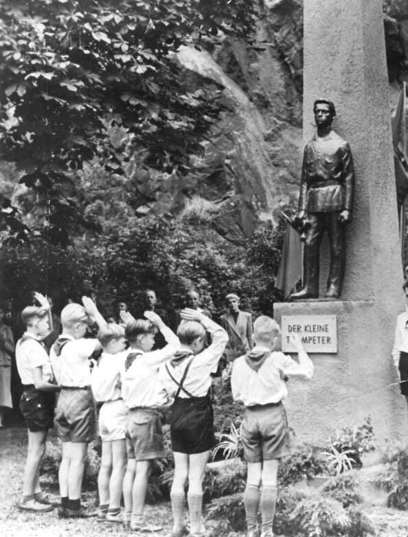 Das schwarz-weiße Foto zeigt eine Gruppe von Jungen in kurzen Hosen, Hemden und Halstüchern, die vor einer Statue mit der Aufschrift „Der kleine Trompeter“ salutieren. Die Statue stellt eine männliche Figur in Uniform dar, die ein Musikinstrument in der Hand hält. Sie steht auf einem steinernen Sockel. Die Szenerie befindet sich in einer bewaldeten, felsigen Umgebung mit mehreren Erwachsenen im Hintergrund, die das Geschehen beobachten. Junge Pioniere aus der DDR-Zeit nehmen an einer Gedenkveranstaltung teil.