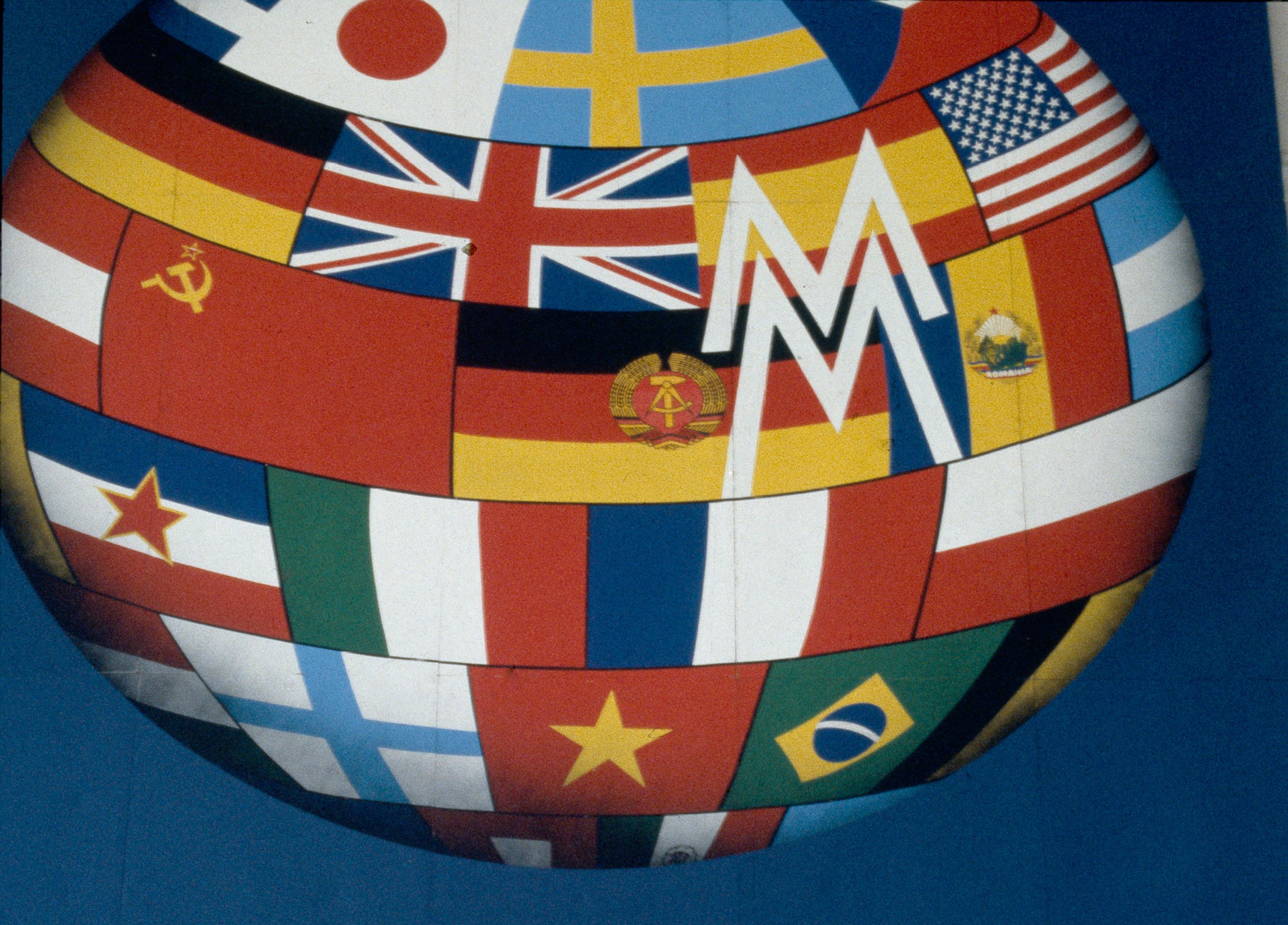 Ausschnitt aus dem Plakat der Leipziger Messe (1989): Eine kugelförmige Oberfläche aus aneinandergesetzten Nationalflaggen füllt das Bild. Über die Flaggen legt sich ein weißes, stilisiertes „M“ als Messezeichen; der Hintergrund ist blau.